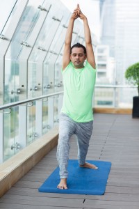 Young Indian man wearing sport clothes and practicing yoga on open terrace of modern fitness club, standing warrior pose with closed eyes. Yoga concept